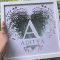 A hand holds a square white laser-cut wooden frame featuring a heart-shaped leafy design, a large letter 'A', and the name 'ADITYA' in a leaf cutout style, set against a background of green palm leaves.