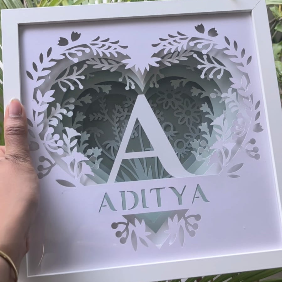 A hand holds a square white laser-cut wooden frame featuring a heart-shaped leafy design, a large letter 'A', and the name 'ADITYA' in a leaf cutout style, set against a background of green palm leaves.